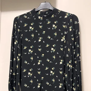 Svart blommig långärmad blus - Svart klänning med små blommor i vitt och gult, långärmad och med hög hals. Tunt och mjukt material som faller snyggt. Perfekt för dig som gillar romantiska och söta detaljer. Blommönstret ger en fräsch och ungdomlig vibe.