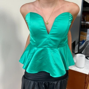 Grön axelbandslös peplum-topp - Snygg grön axelbandslös topp med peplumvolang och djup v-ringning. Toppens glansiga material ger en lyxig känsla och den sitter tight upptill med utsvängd nederdel. 