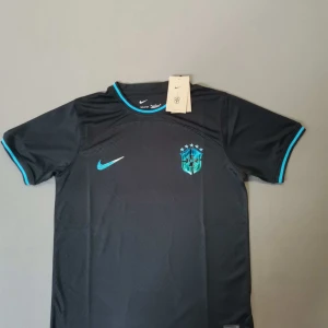 Svart & ljus blå Brasil Nike fotbollströja M - Snygg svart Brasil fotbollströja från Nike med turkosa detaljer på krage och ärmslut. Tröjan har korta ärmar och Brasil-märket på bröstet, samt Nike-logga i turkos. Tillverkad i lätt och ventilerande polyester, perfekt för träning eller match.