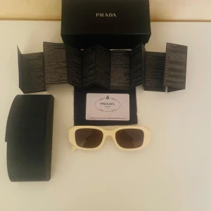 Beige solglasögon från Prada - Snygga solglasögon från Prada med rektangulär form och beige bågar i plast. Mörka linser med diskret Prada-logga på glaset och på skalmen. Kommer med originalfodral, putsduk och äkthetsbevis. Perfekt för dig som vill ha en lyxig och trendig look.
