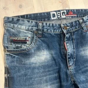 Blå DSQUARED2 jeans med detaljer - Säljer ett par blå jeans från DSQUARED2 med coola slitningar och kontrastsömmar. Jeansen har broderade och patchade detaljer på fickorna, samt röda inslag vid dragkedjan och bälteshällorna. Klassisk femficksmodell i mjukt denimtyg.