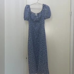 Blommig blå långklänning med puffärm - Säljer en superfin blå långklänning med vita blommor och puffiga kortärmar. Klänningen har en söt, rynkad urringning och dragkedja i ryggen. Perfekt för dig som gillar romantisk stil och vill ha något luftigt och somrigt.