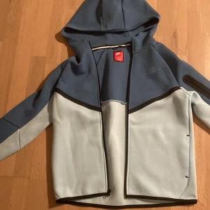 Nike blå och ljusgrå vindjacka XL - Nike vindjacka i blått och ljusgrått med huva och hel dragkedja. Jackan har svarta detaljer, en ficka med dragkedja på ärmen och sportig design. Materialet är mjukt och syntetiskt, perfekt för chill och streetwear. Snygg logga på bröstet och bak.