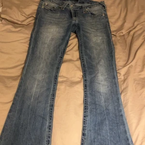 True Religion bootcut jeans blå, stl 28 - Säljer ett par klassiska blå jeans från True Religion i bootcut-modell. Jeansen har låg midja, slitna detaljer och fransade benslut för en cool vibe. Materialet är robust denim med snygga kontrastsömmar och ikoniska bakfickor med broderi. Jeansen är även lite slitna där nere vid benen köpte för 1399