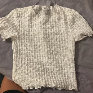 Vit ribbad t-shirt från AWARE XS - Säljer en vit t-shirt från AWARE i storlek XS. T-shirten har korta ärmar och är gjord i ett ribbat, stretchigt material med vågiga kanter vid hals och nederkant. Perfekt för dig som gillar en unik och cool textur på dina kläder.