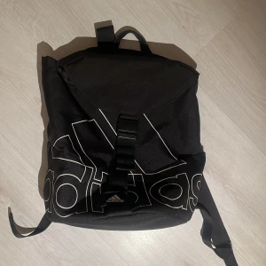 Svart Adidas ryggsäck med logga - Snygg svart ryggsäck från Adidas med stor vit logga framtill. Väskan har justerbara axelremmar, ett stort huvudfack med dragkedja och innerficka. Materialet är slitstark polyester och designen är stilren med en cool sportig vibe. 