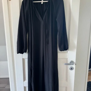 Svart långklänning med v-ringning hm  - Snygg svart långklänning i storlek XS med v-ringad hals och långa ärmar. Klänningen har ett diskret strukturmönster i tyget och knytband vid halsen. Perfekt för dig som gillar enkel och stilren design med lite extra detaljer.