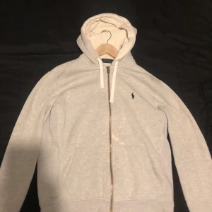 Grå hoodie från Polo Ralph Lauren - En grå hoodie från Polo Ralph Lauren med dragkedja och huva med vita snören. Tröjan har en fläck vid dragkedjan som knappt syns och en vid armen som man inte lägger märke till som kan färgas med kläder färg.
