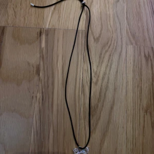 Svart halsband med silverfärgad fjäril - Coolt svart halsband med en detaljrik silverfärgad fjäril som hänge. Halsbandet har justerbar längd med metalländar och en liten silverfärgad pärla vid knytet. Perfekt för dig som gillar unika smycken med naturinspirerade detaljer.