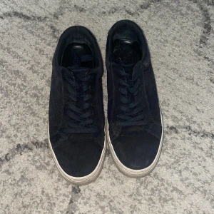 Mörkblå mocka sneakers med vit sula - Snygga mörkblå sneakers med ovandel i mocka och klassisk vit platt sula. Skorna har rund tå och snörning framtill för en clean look. Perfekta för dig som gillar stilrena och enkla sneakers med lite extra känsla tack vare mockamaterialet.