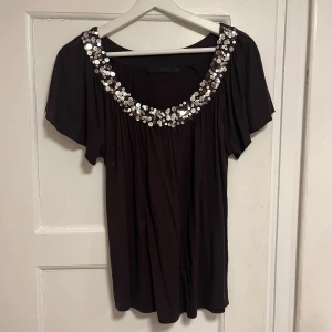 Brun topp med paljetter från Oui Set - Brun topp från Oui Set med korta ärmar och rundad halsringning dekorerad med glittriga paljetter. Toppens material är mjukt och faller snyggt, perfekt för dig som gillar att sticka ut med lite extra bling. Snyggt flowy fit och passar dig som gillar detaljer.