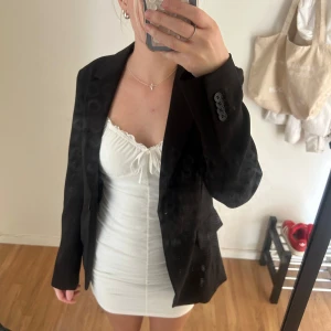 Svart kavaj från H&M - Snygg svart kavaj från H&M i storlek 34. Klassisk modell med krage, knappar vid ärmslut och fickdetaljer framtill. Perfekt att slänga över en klänning eller till jeans för en clean look. Materialet är mjukt och har en lätt glansig finish.