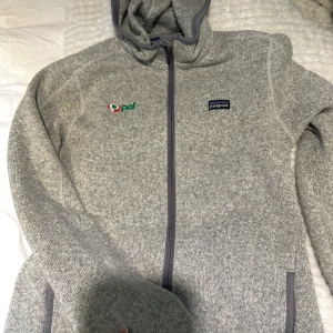 Grå hoodie från Patagonia med dragkedja - Säljer en grå hoodie från Patagonia i mjuk polyester med hel dragkedja och huva. Tröjan har broderade loggor på bröstet och ribbade muddar. Perfekt för kyliga dagar och har en clean, sportig look.