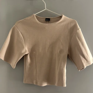 Beige croppad topp från Gina Tricot - Snygg beige croppad topp från Gina Tricot med korta ärmar och rund halsringning. Enkel och stilren design som passar perfekt till jeans eller kjol. Mjuk bomullskänsla och lätt figursydd passform för en modern look.