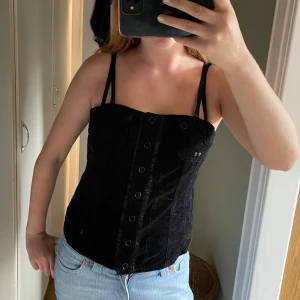 Svart manchesterkorsett med knappar - Säljer en svart korsett-topp i mjuk manchester med dubbla smala axelband och dekorativa knappar framtill. Toppen har en liten ficka på bröstet och är figurnära med coola sömdetaljer. Perfekt att styla med jeans för en edgy look.