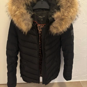 Svart pufferjacka Hollies med päls - Svart pufferjacka från Hollies med fluffig beige päls runt huvan och leopardmönstrat foder. Jackan har dragkedja framtill, två fickor med dragkedja.