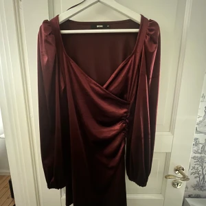 Vinröd omlottklänning från Bik Bok - Snygg vinröd kortklänning från Bik Bok i glansig polyester med omlottdetalj och puffiga långärmar. Klänningen har lätt rynkning i sidan och en v-ringad hals. Perfekt för dig som vill sticka ut med en elegant och trendig look. Passar perfekt till jul! 