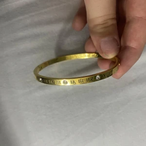 Guldigt armband med romerska siffror - Snyggt och stilrent armband i guldfärgad metall med ingraverade romerska siffror runt hela bandet. Armbandet har flera glittriga stenar som ger extra bling och en lyxig känsla. Perfekt för dig som gillar detaljer och vill sticka ut lite extra. Helt oanvänd! Fått i present