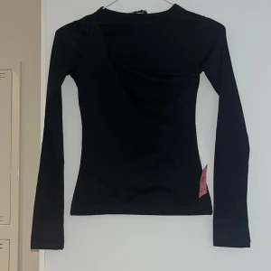 Svart omlott långärmad topp PLT - Svart långärmad topp från PrettyLittleThing med cool omlott-detalj framtill. Slim fit och mjukt bomullsmaterial som sitter snyggt mot kroppen. Perfekt för dig som gillar stilrena plagg med en twist. Passar till jeans eller kjol för en trendig look.