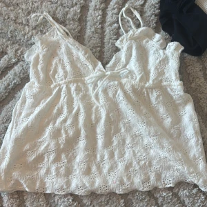 Vit spetsig linnetopp med knyt - Supersöt vit linnetopp med tunna axelband och v-ringning framtill. Toppen har broderade spetsdetaljer och ett knyt under bysten som ger en extra touch. Perfekt för varma dagar och har en luftig, feminin look.