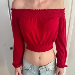 Röd offshoulder croppad topp - Snygg röd offshoulder topp med lång ärm och croppad passform. Smockad kant upptill och nedtill ger en cool och figurnära look. Perfekt att matcha med jeans för en trendig vibe. Materialet känns lätt och bekvämt mot huden.