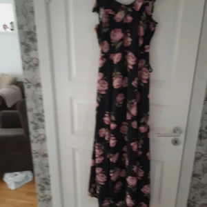 Svart långklänning med rosa blommor - Supersnygg svart långklänning med rosa blommor och gröna blad. Klänningen har omlott-detalj i ryggen, knytband och dragkedja i sidan. Ärmlös modell med mjukt fallande tyg, perfekt för sommarkvällar. Blommönstret ger en romantisk vibe och klänningen sitter normal i passform.