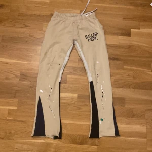 Beige Gallery Dept. mjukisbyxor S - Unika beige mjukisbyxor från Gallery Dept. med svart textprint på framsidan och konstnärliga färgstänk i vitt, svart och grönt. Byxorna har svarta paneler längs bensluten, elastisk midja med snörning och är tillverkade i 100% bomull. Perfekt för dig som gillar streetwear med edge.