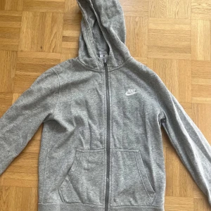 Grå hoodie från Nike med dragkedja - Grå hoodie från Nike med klassisk logga på bröstet. Tröjan har huva, långa ärmar och praktiska fickor framtill. Materialet är mjuk bomull och polyester, perfekt för chill eller träning. Dragkedja hela vägen och ribbade muddar för extra komfort. Är storlek L för barn men skulle säga att det är runt xs. Skriv vid minsta fråga!💕💕