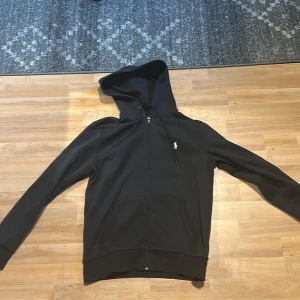 Svart hoodie från Polo Ralph Lauren - Svart hoodie från Polo Ralph Lauren med dragkedja och huva. Klassisk design med logotyp på bröstet. Använd få tal gånger 