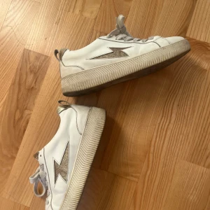 Vita sneakers från Vanessa wu - Snygga vita sneakers från W The Designer med glittrig blixtdetalj på sidan och guldfärgade detaljer på plösen och hälen. Skorna har rund tå, platt sula och klassisk snörning. Perfekta för dig som gillar stilrena men ändå unika sneakers.