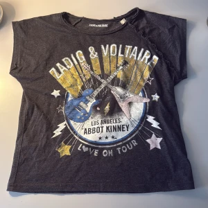 Svart t-shirt Zadig & Voltaire med tryck - Cool svart t-shirt från Zadig & Voltaire. Storleken är 138 men kan även bäras på långre personer än det då den kan bli lite mer kort/croppad.  märgstarkt tryck av gitarrer, stjärnor och texten 'Love on Tour'. T-shirten har korta ärmar, rund hals och snygga knappar längs ena axeln. Materialet är mjuk bomull. Svart/grå.