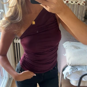 Vinröd one shoulder topp från Bik Bok - Säljer en vinröd one shoulder topp från Bik Bok. Toppen är tight och har snygga rynkade detaljer längs sidorna. Gjord i ett stretchigt, glansigt syntetmaterial som sitter skönt mot kroppen. Perfekt för dig som vill sticka ut med en cool och trendig look.