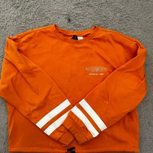 Orange sweatshirt Sacramento 1991 - Cool orange sweatshirt från H&M med trycket 'Sacramento California 1991' på bröstet. Långärmad med vita ränder på ärmarna och snörning i nederkant för extra stil. Perfekt för dig som gillar streetwear och vill sticka ut med färg.