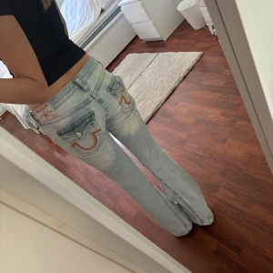 Skitsnygga True Religion ljusblå bootcut jeans - Säljer ett par ikoniska ljusblå bootcut jeans från True Religion med fickor bak med deras klassiska logga. Jeansen har låg midja och är tillverkade i mjukt denim med snygg tvättad look. Perfekta, använda 1 gång men för stora. För referens är jag 170cm och har storlek xs/s, dessa är storlek 26 och modell Joey. Pris kan diskuteras vid snabb affär.🥰