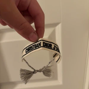 Christian Dior J'adore armband - Snyggt armband från Christian Dior med texten 'J'adore' broderad i svart på en vit bakgrund. Armbandet är gjort i ett mjukt tygmaterial och har coola tofsar i ändarna som knyts runt handleden. Perfekt accessoar för att lyfta din outfit med lite lyxig vibe.
