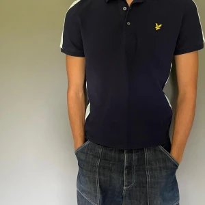 Mörkblå pikétröja från Lyle & Scott - Snygg mörkblå pikétröja från Lyle & Scott med korta ärmar och ljusblå och vita ränder på sidorna.               Skick 8/10