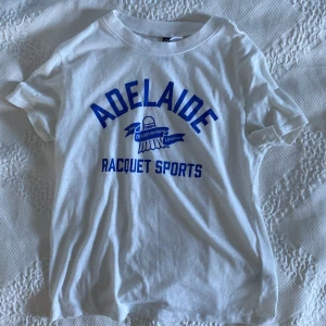 Vit t-shirt H&M XS - Vit t-shirt från H&M Divided i storlek XS med blått tryck framtill: 'ADELAIDE RACQUET SPORTS' och en badmintonikon. Rund hals och korta ärmar. Perfekt för dig som gillar sportig och chill stil.