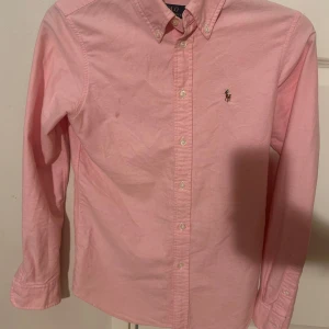 Rosa skjorta från Polo Ralph Lauren XS - Snygg rosa skjorta från Polo Ralph Lauren i bomull, custom fit. Klassisk button-down krage, broderad logga på bröstet och vita knappar hela vägen. Långärmad modell med stilren look, perfekt för dig som gillar preppy stil.