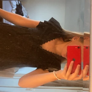 Svart blus med spets och volang - Supersöt svart blus med v-ringad spetskant och volang vid axlarna. Blusen har korta ärmar och är gjord i ett bomullstyg med broderade hål för en extra cool look. Perfekt att styla med jeans eller kjol för en trendig vibe.