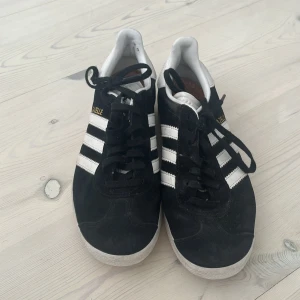 Adidas Gazelle svarta sneakers - Svarta Adidas Gazelle sneakers med klassiska vita ränder och gul logga på sidan. Skorna har snörning, rund tå och platt sula. Ovandelen är i mocka och detaljer i vitt skinn. Perfekt för dig som gillar retrostil och vill ha en ikoniska sneakers i garderoben.