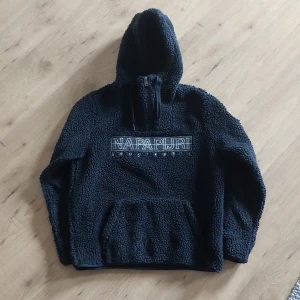 Svart fleece hoodie från Napapijri - Mysig svart hoodie från Napapijri med broderad logga på bröstet. Tröjan är tillverkad i fluffig fleece, har huva, lång ärm och en stor magficka. Perfekt för kalla dagar och chill stunder. Half zip framtill för extra stilpoäng.