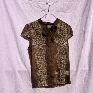 Leopardmönstrad blus från Hillevi - Snygg transparent blus med leopardmönster från Hillevi i storlek 36. Blusen har korta ärmar, rund hals och en cool rosettdetalj framtill. Materialet är 100% polyester och färgerna går i brunt och beige. 