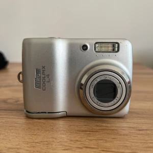 Nikom coolpix digital kamera - Den är köpt här på Vinted. Jag har personligen aldrig använt själv då jag har en annan vilket är anledningen till varför jag säljer denna. Allt verkar vara okej den sätts på normalt. Batteri ingår men tyvärr inget minneskort då jag inte har ett som passar den. Batteriluckan är tyvärr sönder så jag rekommenderar att lägga lite tejp där för extra säkerhet så att de inte ramlar ut. Priset kan diskuteras