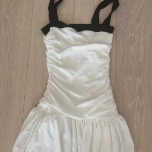 Vit  kortklänning Gina Tricot - Supersöt vit klänning från Gina Tricot i storlek XS med svarta axelband och kant upptill. Klänningen har en rynkad överdel och ett lätt utställt kjolparti. Perfekt för dig som gillar stilrena kontraster och vill ha en enkel men snygg look.