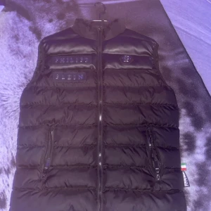 Svart dunväst från Philipp Plein - Svart dunväst från Philipp Plein med coola logopatchar på bröstet och en snygg quiltad design. Västen har två dragkedjefickor och hög krage. Materialet är polyester och dun, med detaljer i syntet. Perfekt för dig som gillar streetwear och vill sticka ut.