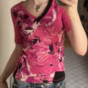 Rosa blommig v-ringad topp - Säljer en rosa v-ringad topp med blommigt mönster i svart och ljusrosa. Toppen har korta ärmar och en cool svart detalj vid halsringningen. Perfekt för dig som gillar färg och vill sticka ut lite extra.