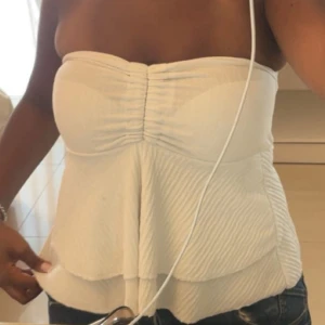 Vit bandeau tubtopp med volang - Supersöt vit bandeau tubtopp med volangdetaljer och lätt rynkning framtill. Toppen är axelbandslös och har en flowy, luftig känsla som passar perfekt till jeans eller kjol. Enkel och stilren design som ger en fräsch look.