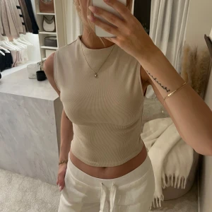Beige ärmlös topp från Zara - Säljer en stilren beige ärmlös topp från Zara. Perfekt för lager-på-lager eller att bära som den är. Toppen har en mjuk och ribbad textur som ger en skön känsla.