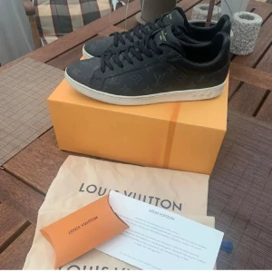 Svarta sneakers från Louis Vuitton - Säljer ett par svarta Louis Vuitton sneakers med diskret LV-mönster över hela skon. Skorna har snörning, rund tå och platt sula. Klassisk och stilren design med märkets logga på sidan och tungan.