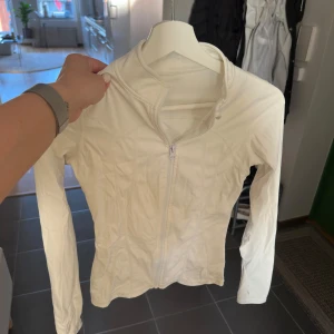 Vit träningsjacka - Säljer en vit, figurnära träningsjacka med dragkedja framtill och lång ärm. Jackan har en sportig look och är tillverkad i ett stretchigt syntetmaterial som passar perfekt till träning eller aktiva dagar. Stl XS